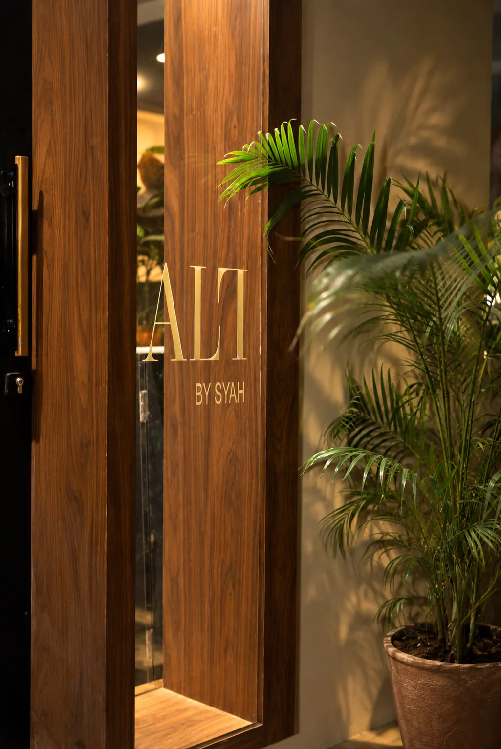 The gold ALT by Syah lettering on the wooden entrance door, beside a potted palm