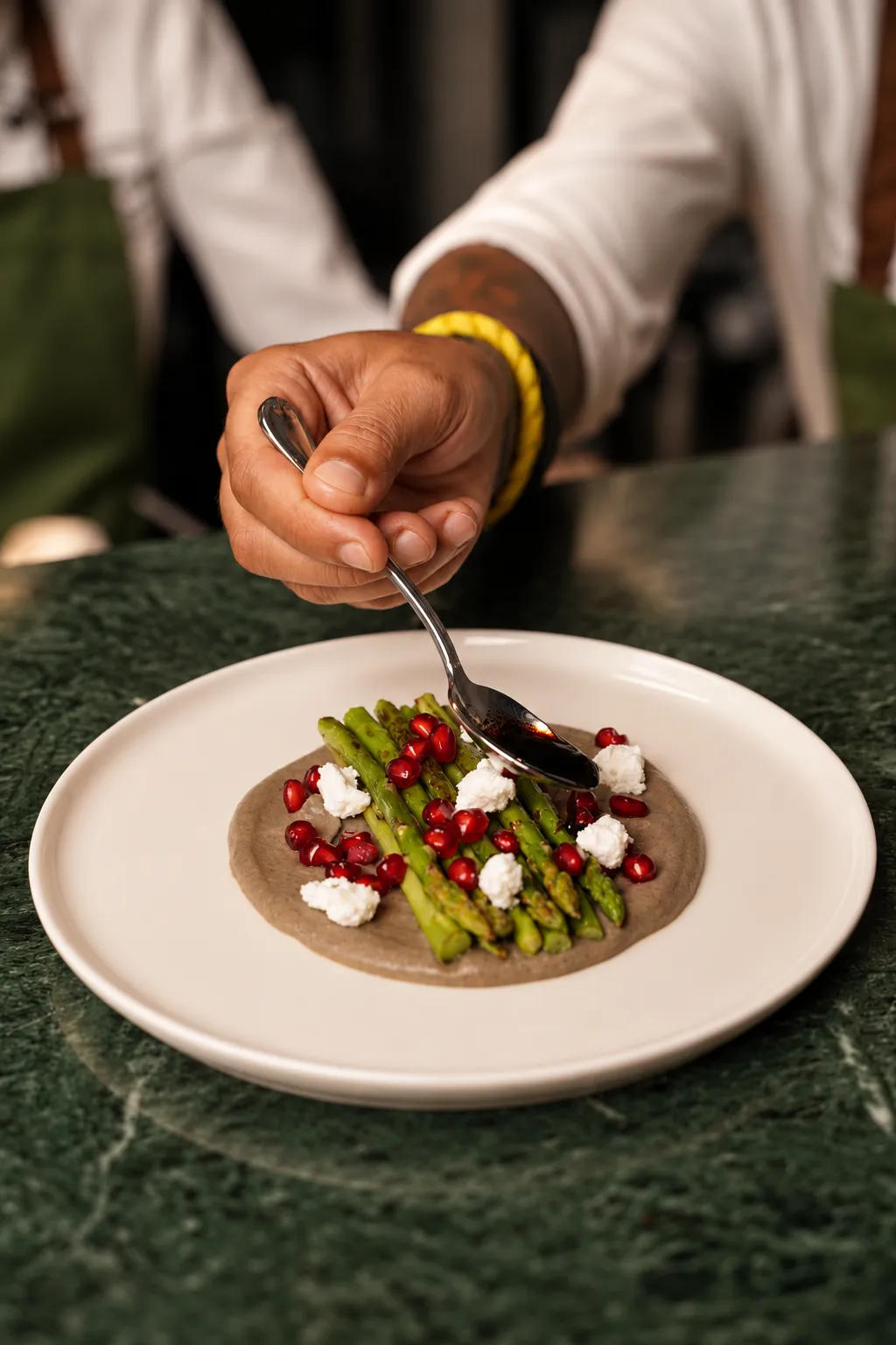 A chef spooning sauce over grilled asparagus with feta and pomegranate